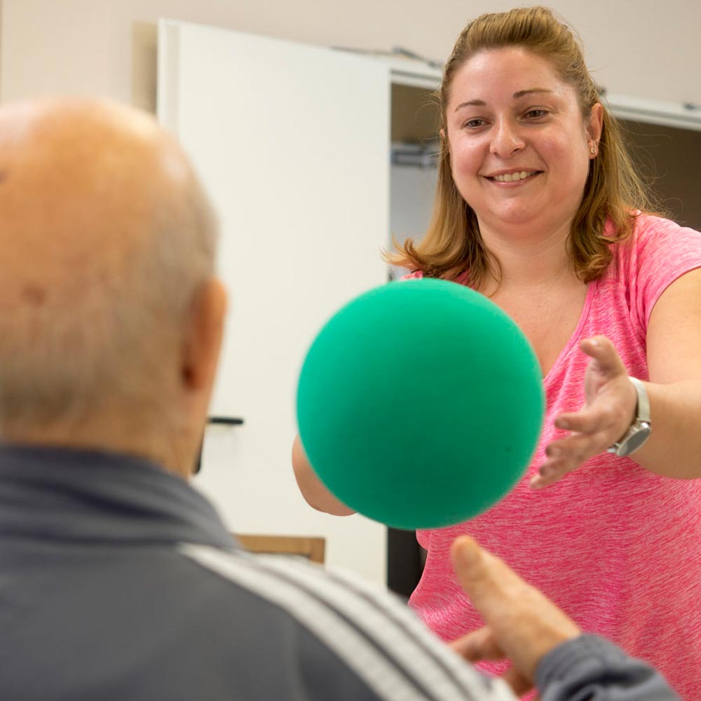 Una trabajadora de la residencia ayuda a un residente en sus ejercicios de actividad física con una pelota